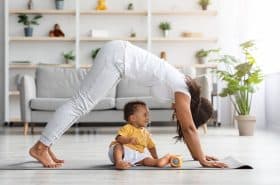 A Black mother wearing a gray shirt and pants doing a downward dog yoga pose over her infant son, who is wearing a yellow shirt and white shorts.